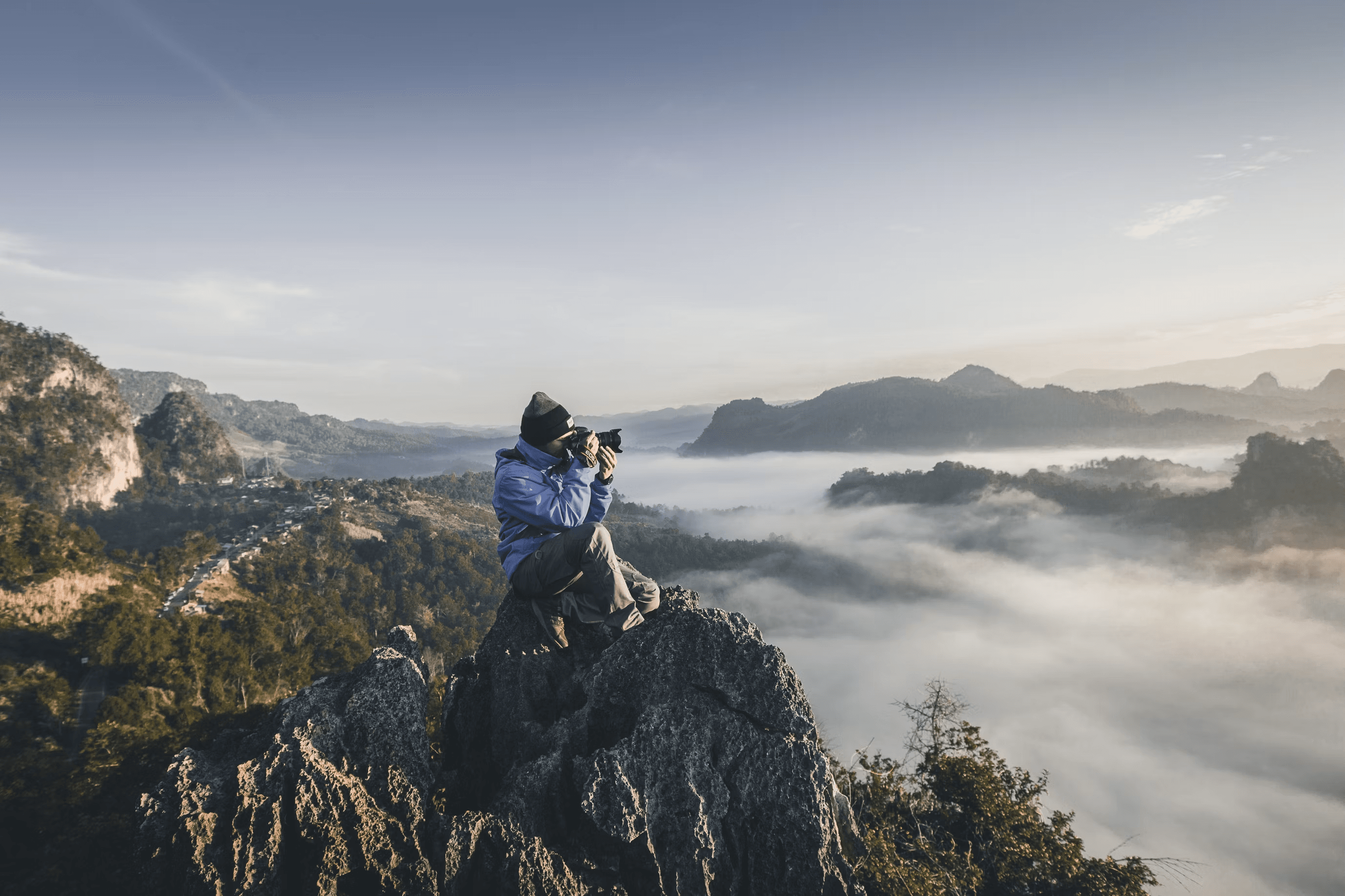 Fotograf i ulur në kreshtën e një mali mbi re, duke fotografuar me një objektiv telefoto